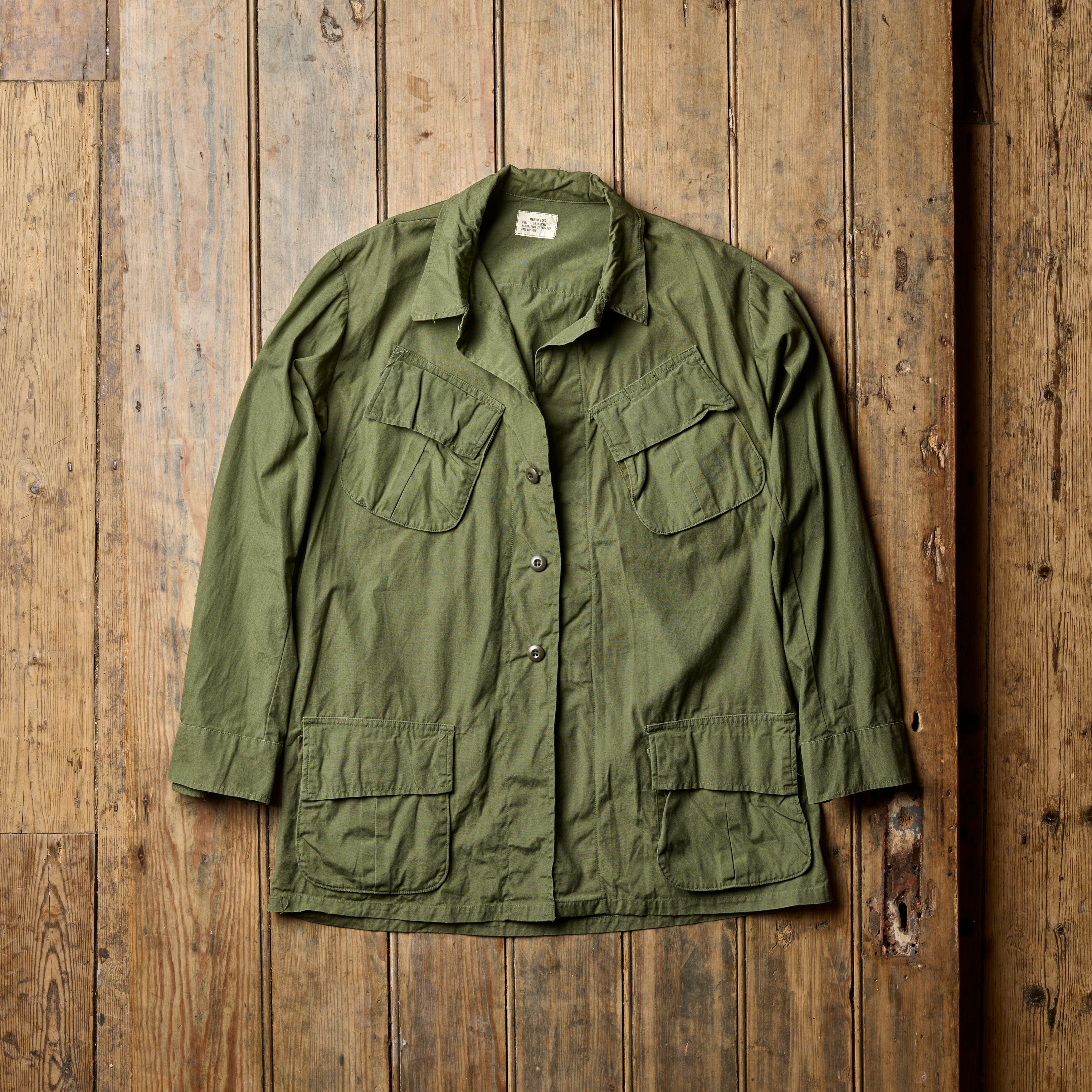 A green U.S. Army Jungle Jacket laid out on a wooden surface, showing the front view with pockets and button details.